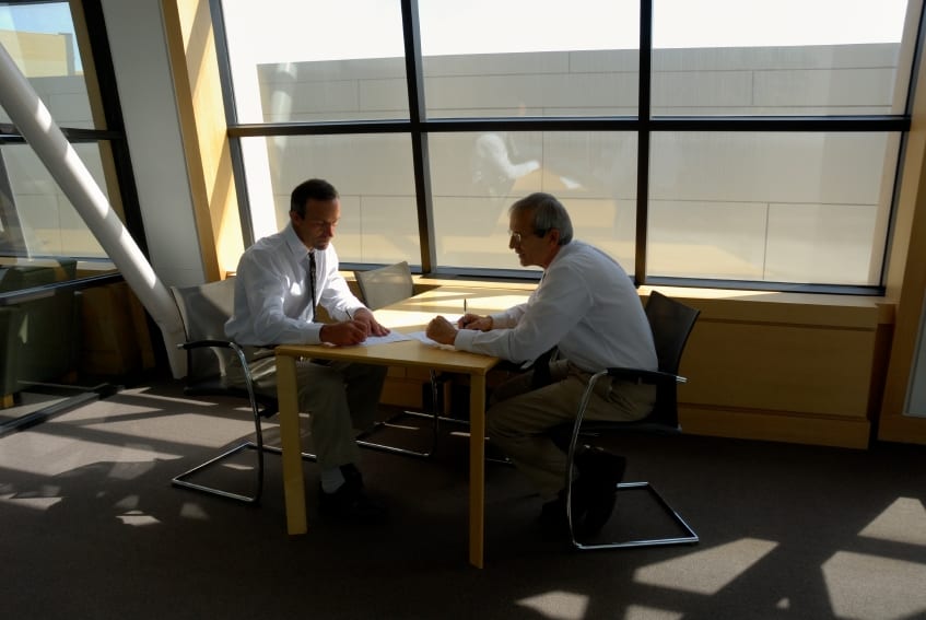 Consulting Photo Two Businessmen Sitting At A Table With Pens In Hand. One Signing The Contract. Big Light Filled Windows Behind Them.